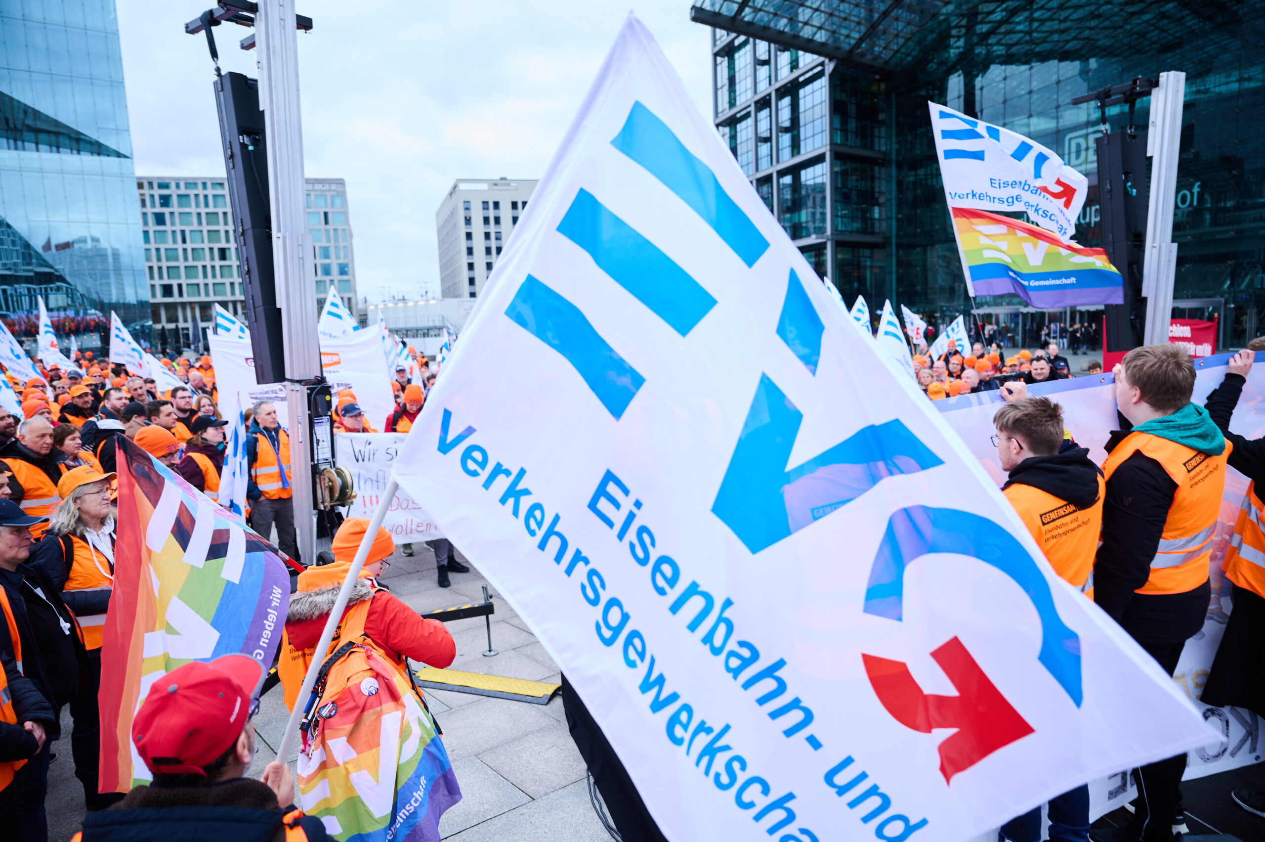 Man sieht eine Menschengruppe vor dem Berliner Hauptbahnhof mit Warnwesten und Flaggen, ganz groß und mittig im Bild ist eine EVG-Fahne.