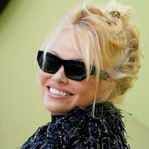 Pamela Anderson lächelt mit Sonnenbrille über ihre Schulter in die Kamera.
