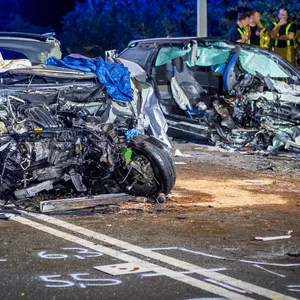 Man sieht zwei Autos, ihre Motorhaube ist eingedrückt, man sieht den Motorrad, teilweise fehlen Stücke der Motorhaube. Dahinter Einsatzkräfte und ein Feuerwehr-Fahrzeug.