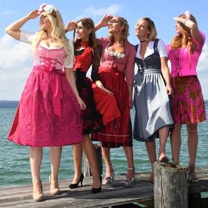 Fünf Frauen in Dirndlkleidern – pink, rot und schwarz-grau – stehen auf einem Steg am See