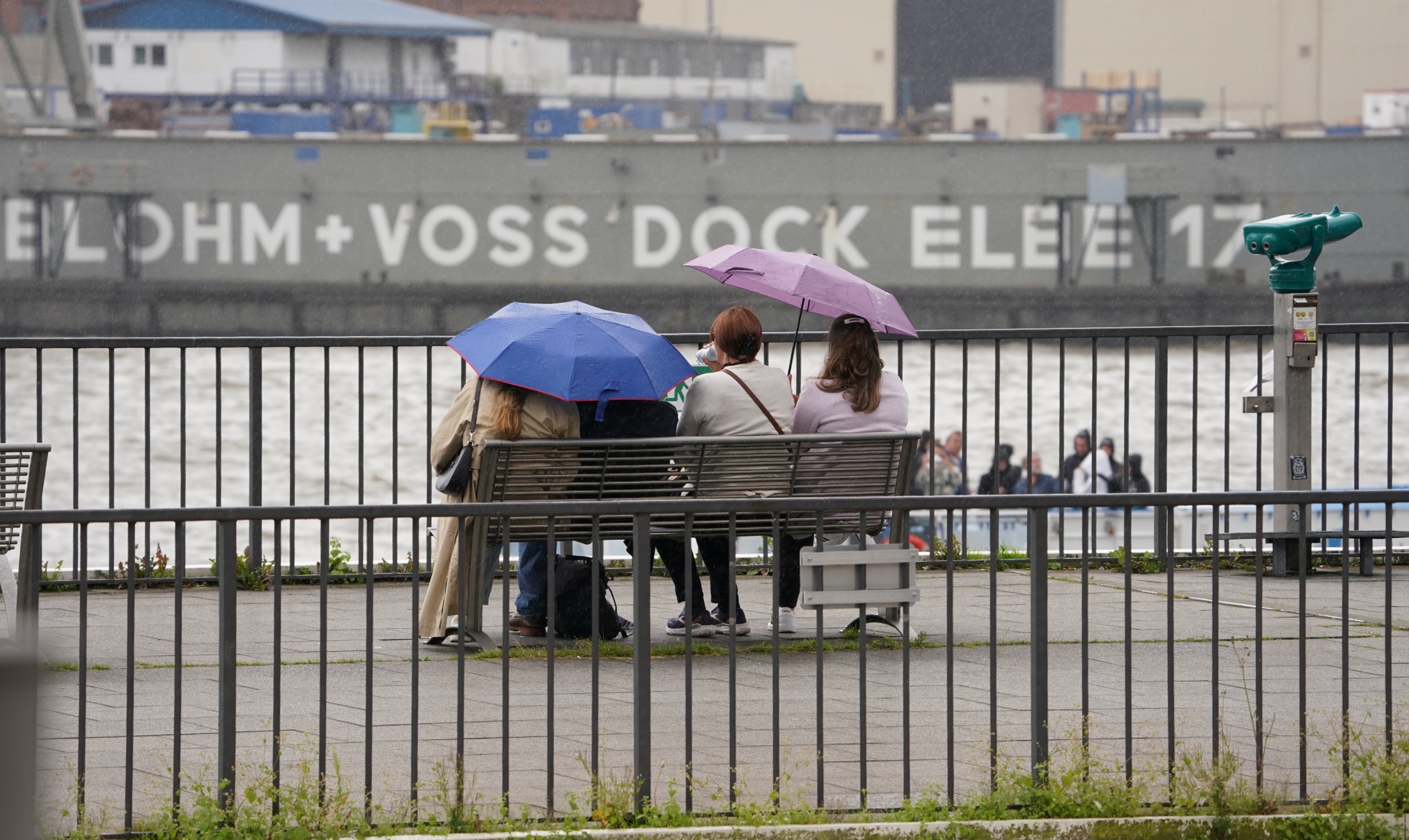 Passanten schützen sich mit Regenschirmen auf einer Sitzbank an den Landungsbrücken vor dem Regen