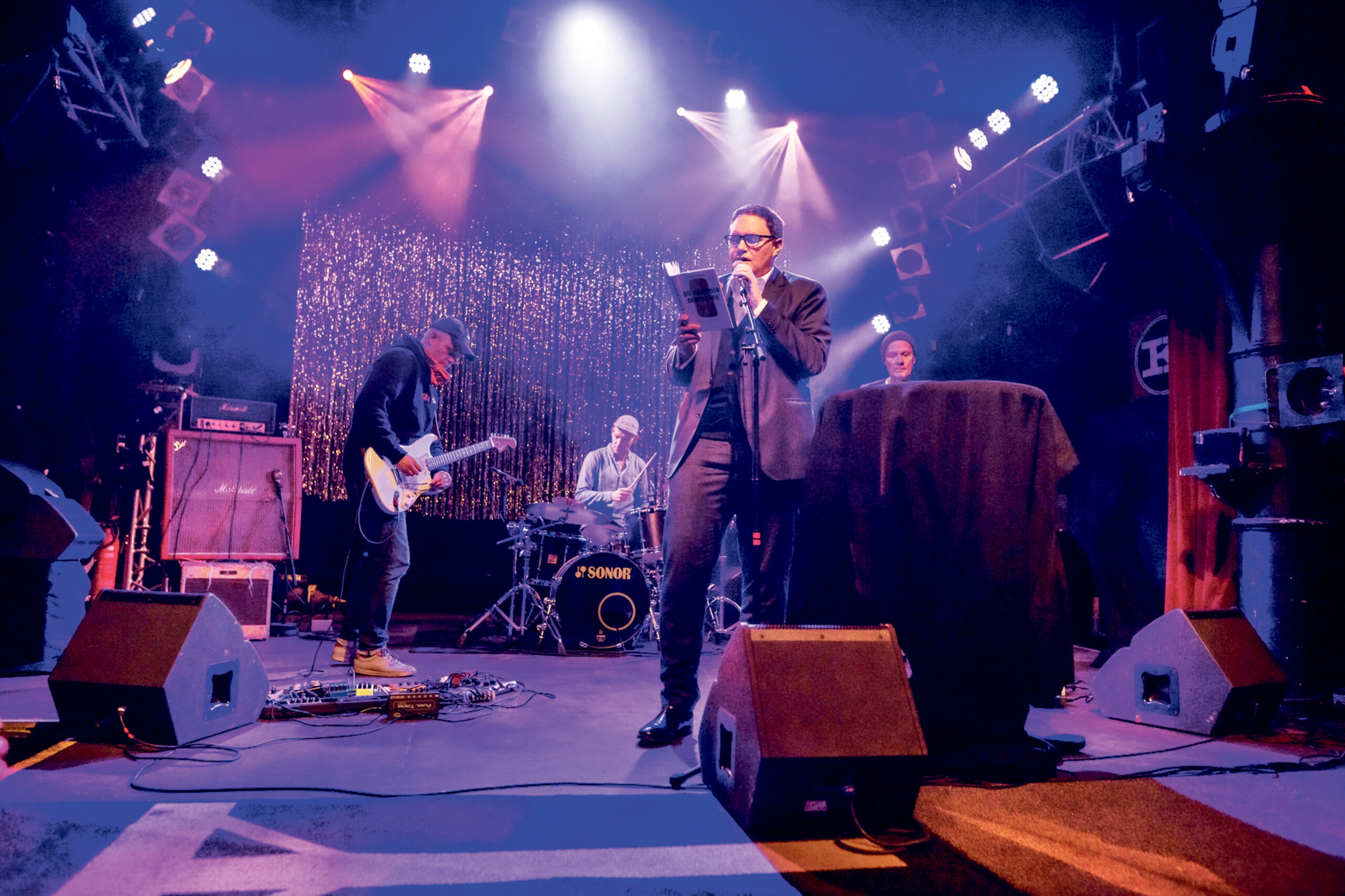 Brosda mit Mikrofon und Buch vorne, im Hintergrund die drei Musiker von Das Weeth Experience