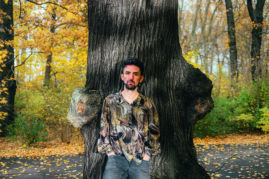 Foto draußen, Thelen lehnt an einem Baum, Hände in den Hosentaschen