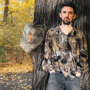 Foto draußen, Thelen lehnt an einem Baum, Hände in den Hosentaschen