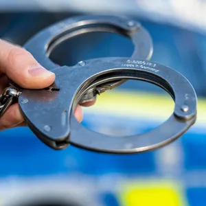Eine Hand hält Handschellen vor einen Streifenwagen der Polizei. (Symbolbild)