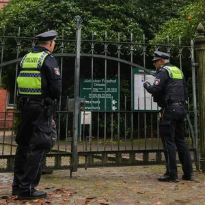 Zwei Streifenbeamte des zuständigen Polizeireviers sprechen am geschlossenen Eingang zum jüdischen Friedhof in Hamburg-Ohlsdorf mit einem Friedhofsmitarbeiter.