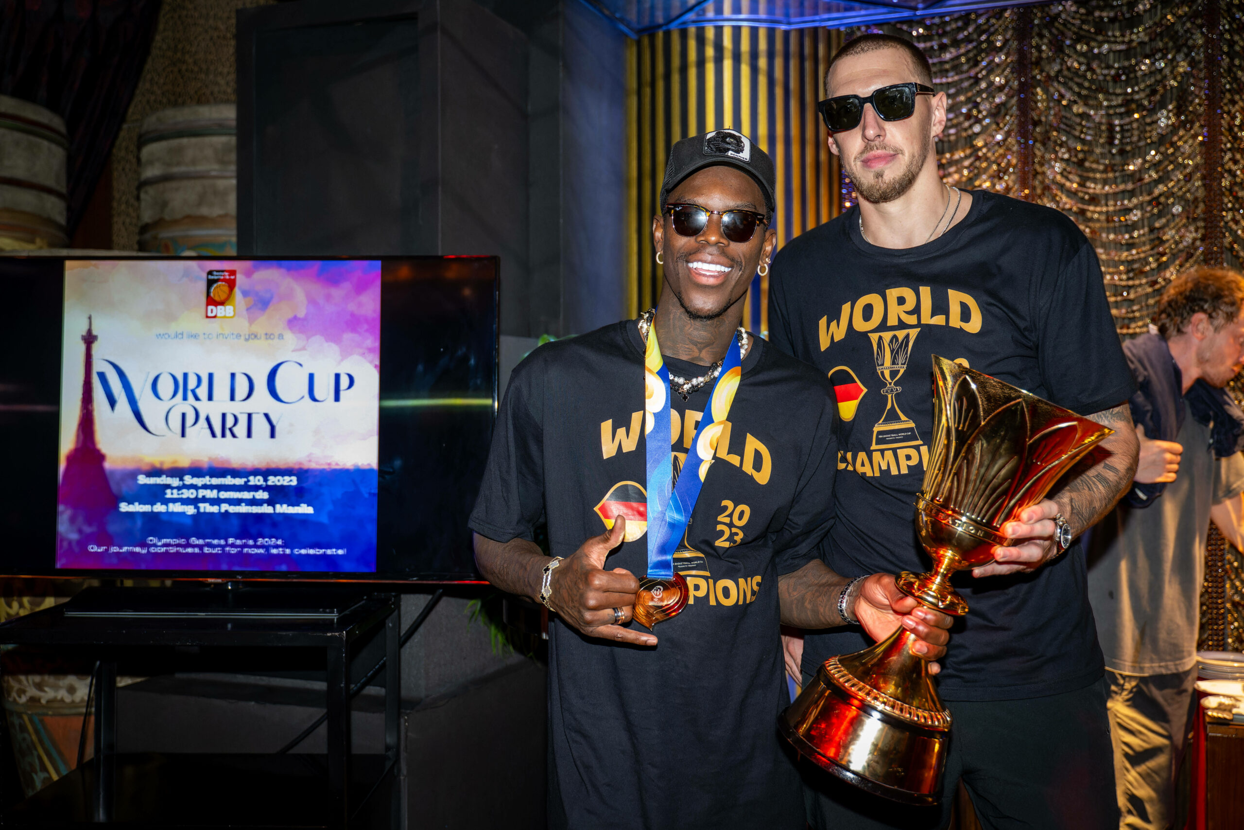 Dennis Schröder und Daniel Theis mit dem WM-Pokal in der Hand, den Sonnenbrillen auf den Augen, lächeln in die Kamera.