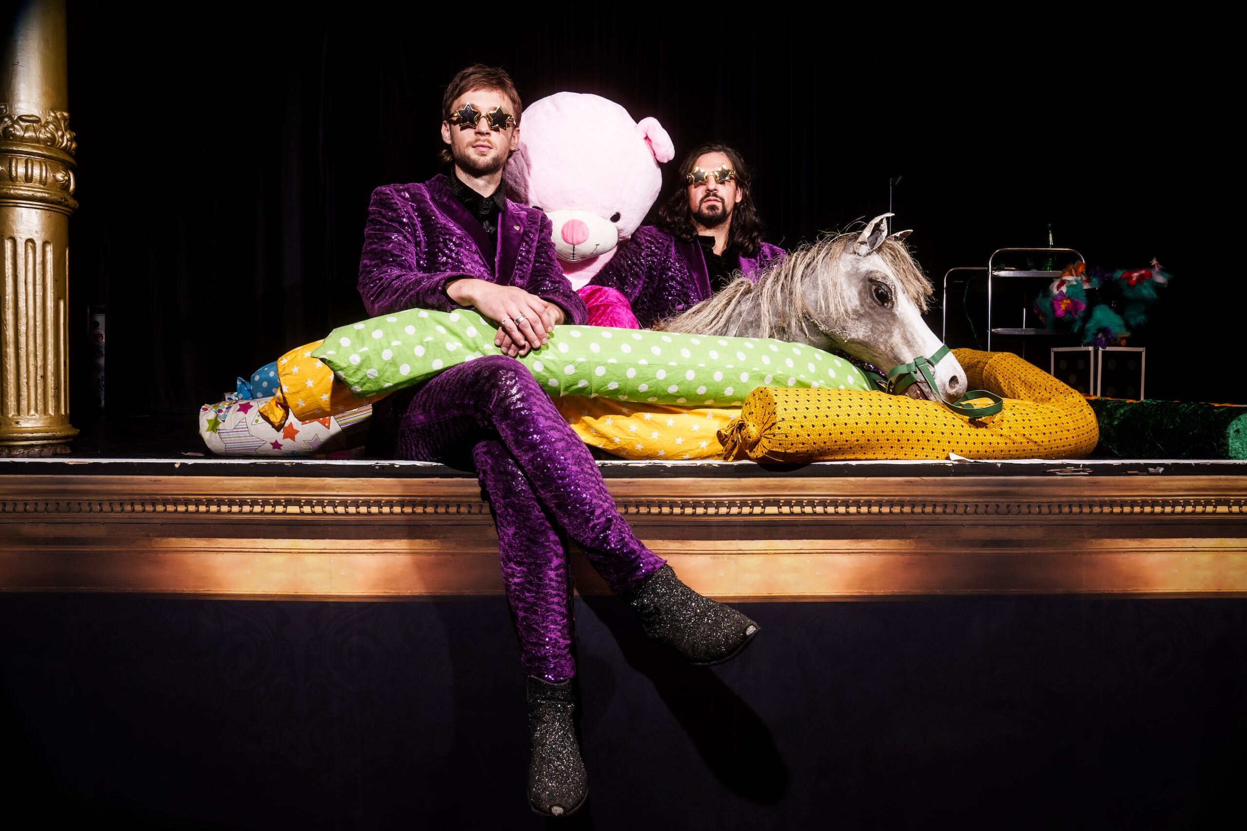 Sitzen in violetten Anzügen und mit Sonnenbrillen auf der Bühne, Kissen, ein großer Stofftier-Bär und ein Pferdekopf zwischen ihnen