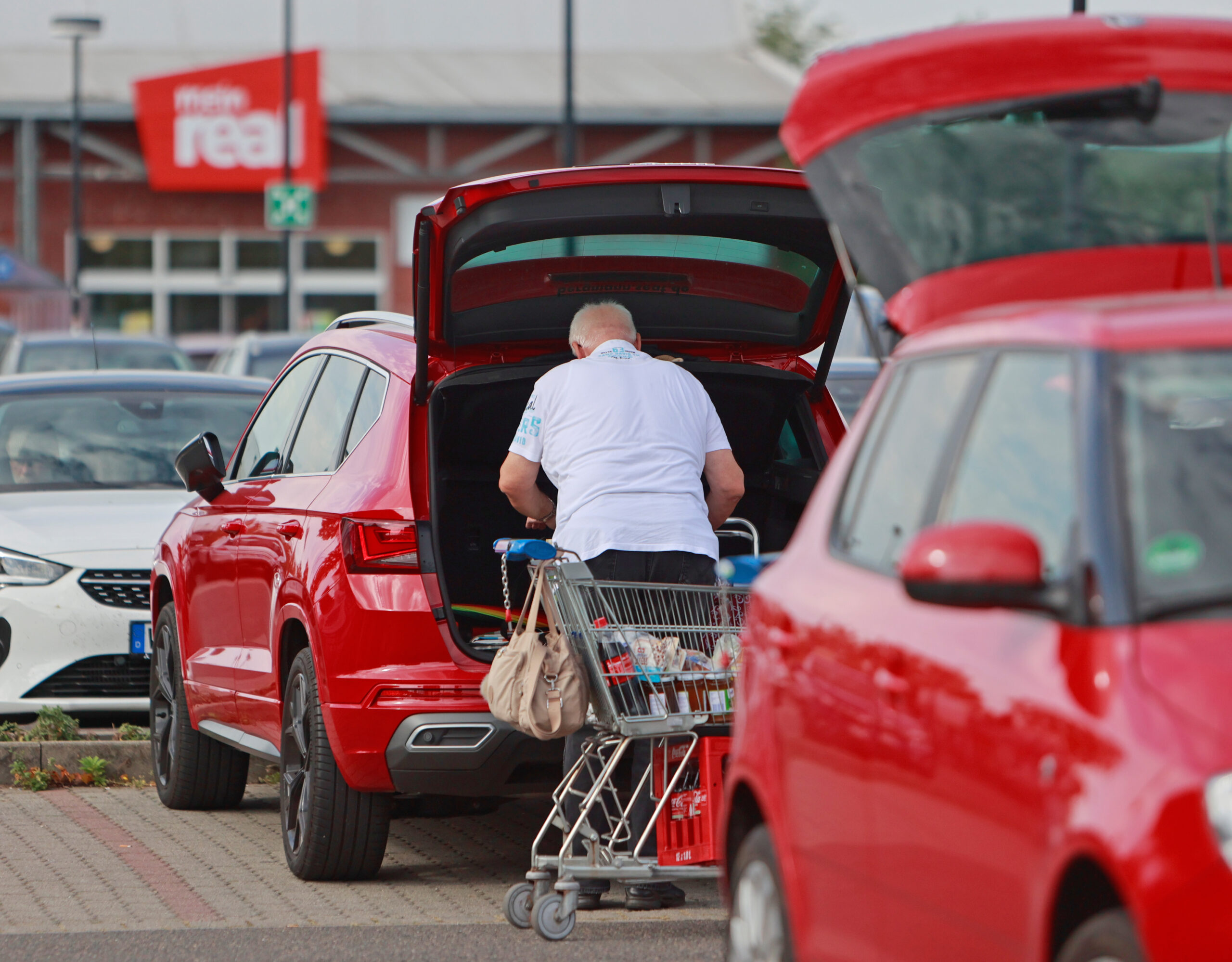Ein Mann steht zwischen zwei geparkten Autos an einem geöffneten Kofferraum, im Hintergrund ist das Logo der SB-Warenhauskette Mein Real zu sehen. (Symbolbild)