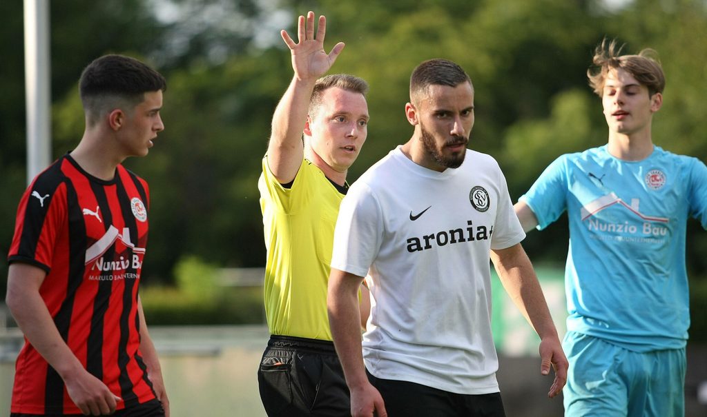 Der Schiedsrichter hebt den Arm und streckt fünf Finger. Elversbergs Alexander (v.r) und Shehada Giuliano Buhtz von Saarbrücken (v.l.) stehen vor ihm