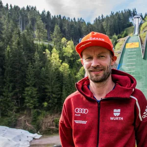 Maximilian Mechler steht vor einer Skisprungschanze.