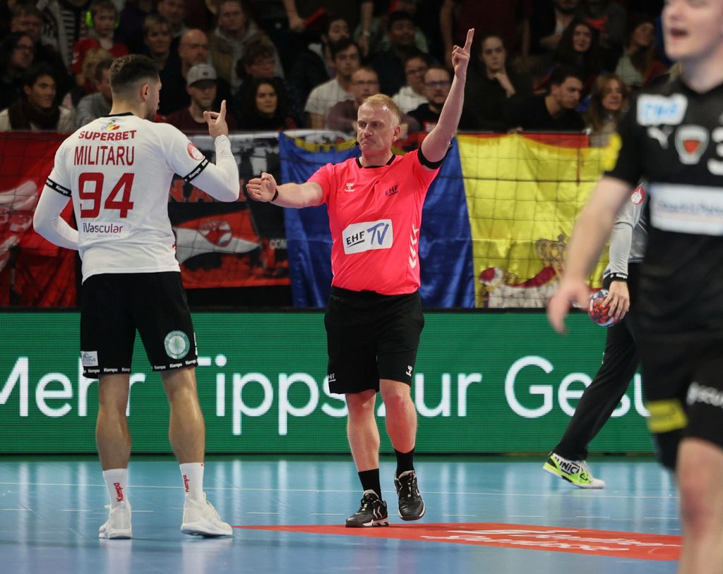 Der Schiedsrichter hebt den linken Arm und streckt zwei Finger nach oben. Den rechten Arm hält er gerade vor den Körper weg, auf den Spieler gerichtet.