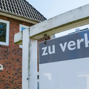 Ein Schild mit der Aufschrift „zu Verkaufen“ steht vor einem Haus. Im Hintergrund scheint die Sonne und es ist ein Baum mit grünen Blättern zu sehen.