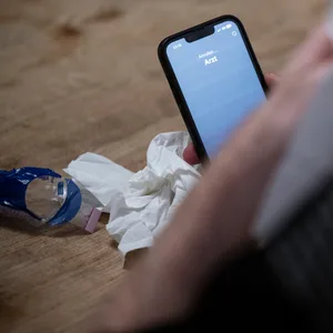 Ein Mann hält ein Taschentuch in einer Hand und ruft mit seinem Smartphone beim Arzt an. (Symbolbild)