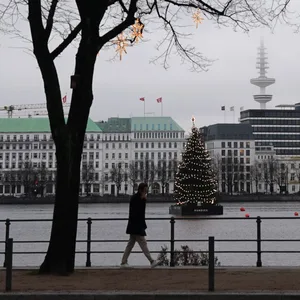 Ein Passant geht an der Binnenalster an weihnachtlich beleuchteten Bäumen entlang. Im Hintergrund leuchtet die Alstertanne.