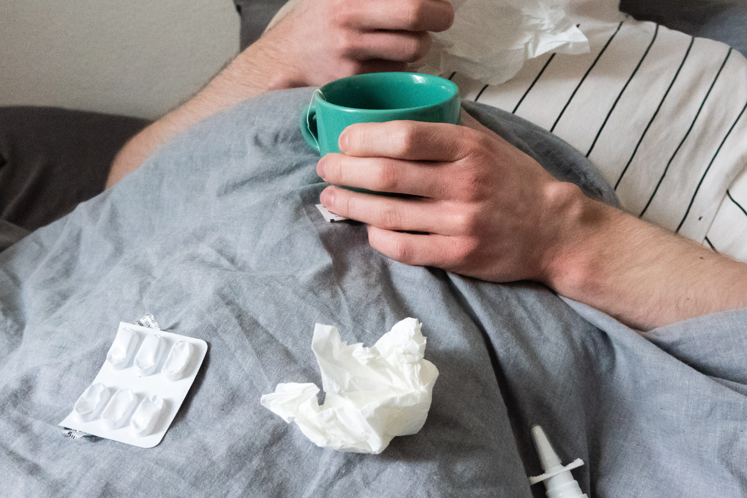 Ein Mann liegt mit Taschentüchern, Teetasse, Nasenspray und Tabletten im Bett. (Symbolbild)
