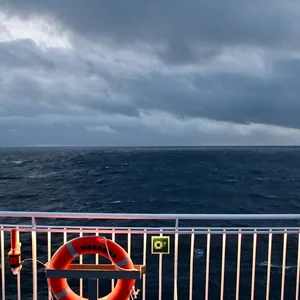 Reling bei Nacht mit Rettungsring, dahinter dunkel das Meer und Wolken