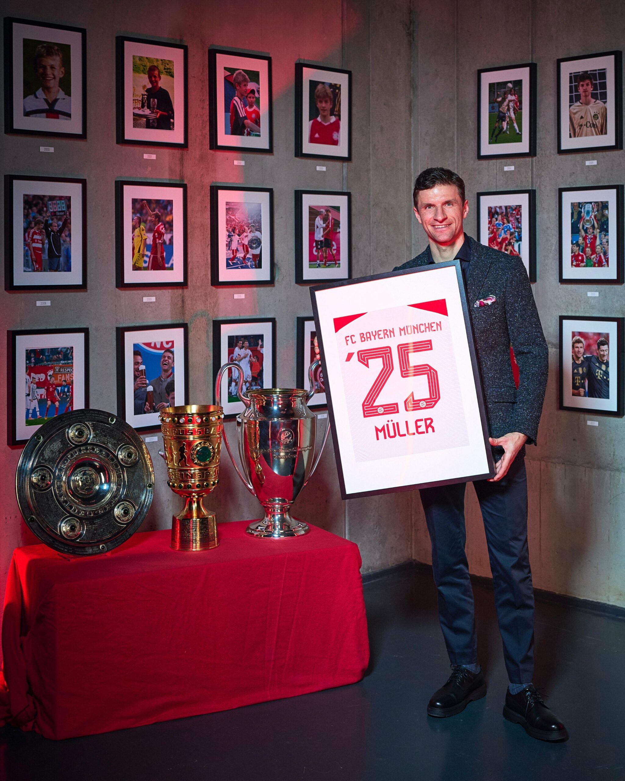 Thomas Müller hält einen Rahmen mit seinem Trikot in der Hand. An der Wand hängen Bilder von Müller, neben ihm steht die Meisterschale, der DFB-Pokal und der Pokal der Champions League