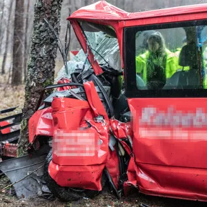 Ein schwer beschädigter kleiner Schultransporter steht am Straßenrand neben einem Baum. Teile des Fahrzeugs wurden durch eine Kollision abgetrennt. Im Hintergrund stehen Rettungsleute.