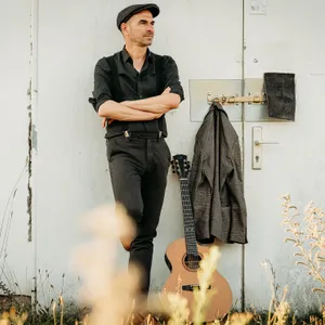 Er lehnt sich draußen an eine Tür, am Türriegel hängt seine Jacke, neben ihm steht seine Gitare