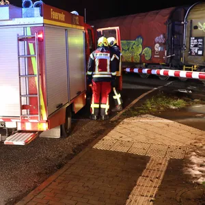 Feuerwehrleute stehen neben einem Rettungswagen, der vor einem Bahnübergang mit roter Ampel steht. Auf dem Gleis steht ein Güterzug, der mit einem Auto kollidiert ist.
