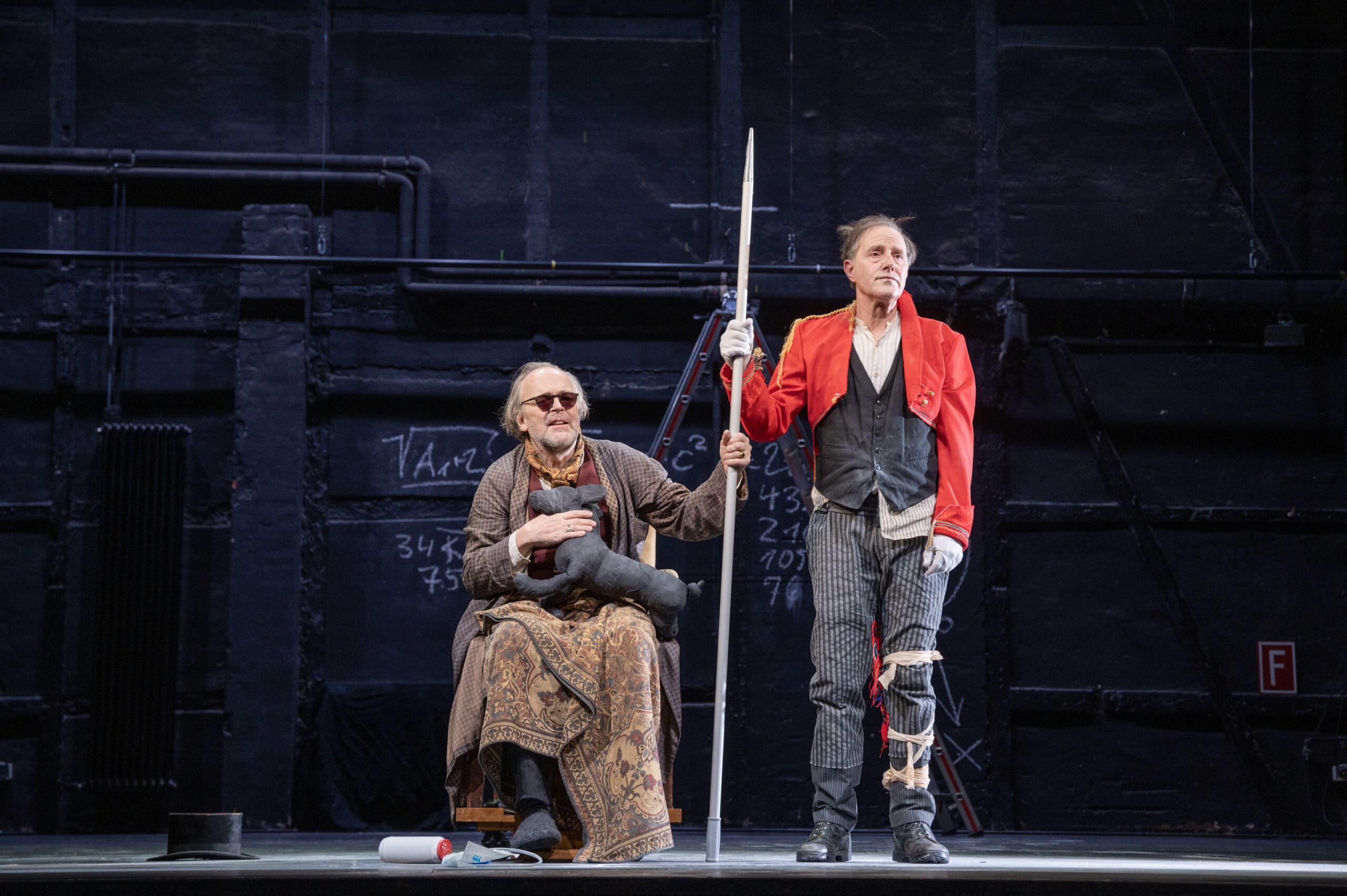 Theaterbühne. Links ein Mann im Rollstuhl und mit Sonnenbrille, rechts ein Mann mit verbundenem Bein. Sie halten sich an einer Stange fest