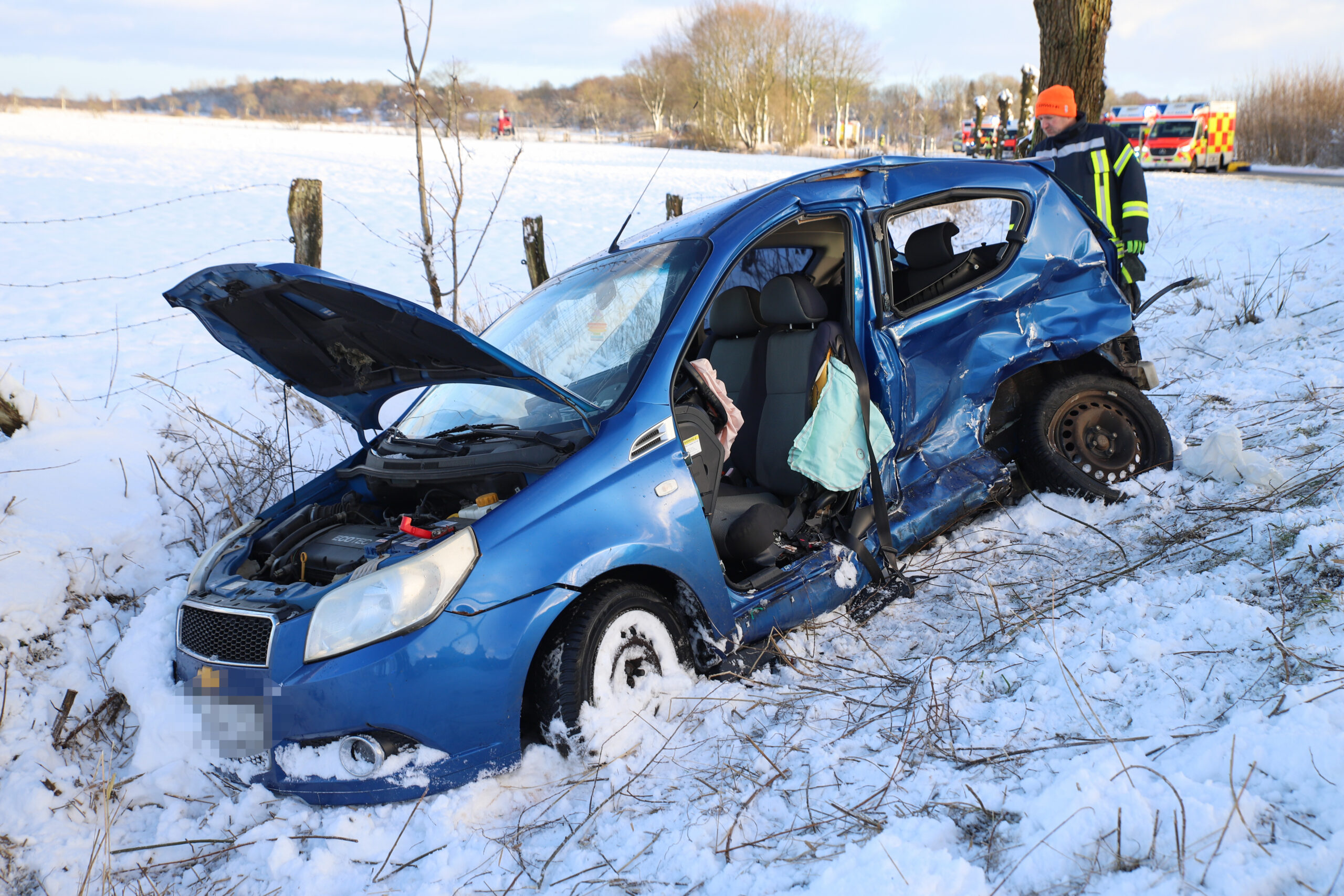 Ein blaues Auto liegt in einem Graben im Schnee. Die Motorhaube steht hoch, eine Tür fehlt. Hinten steht ein Feuerwehrmann.