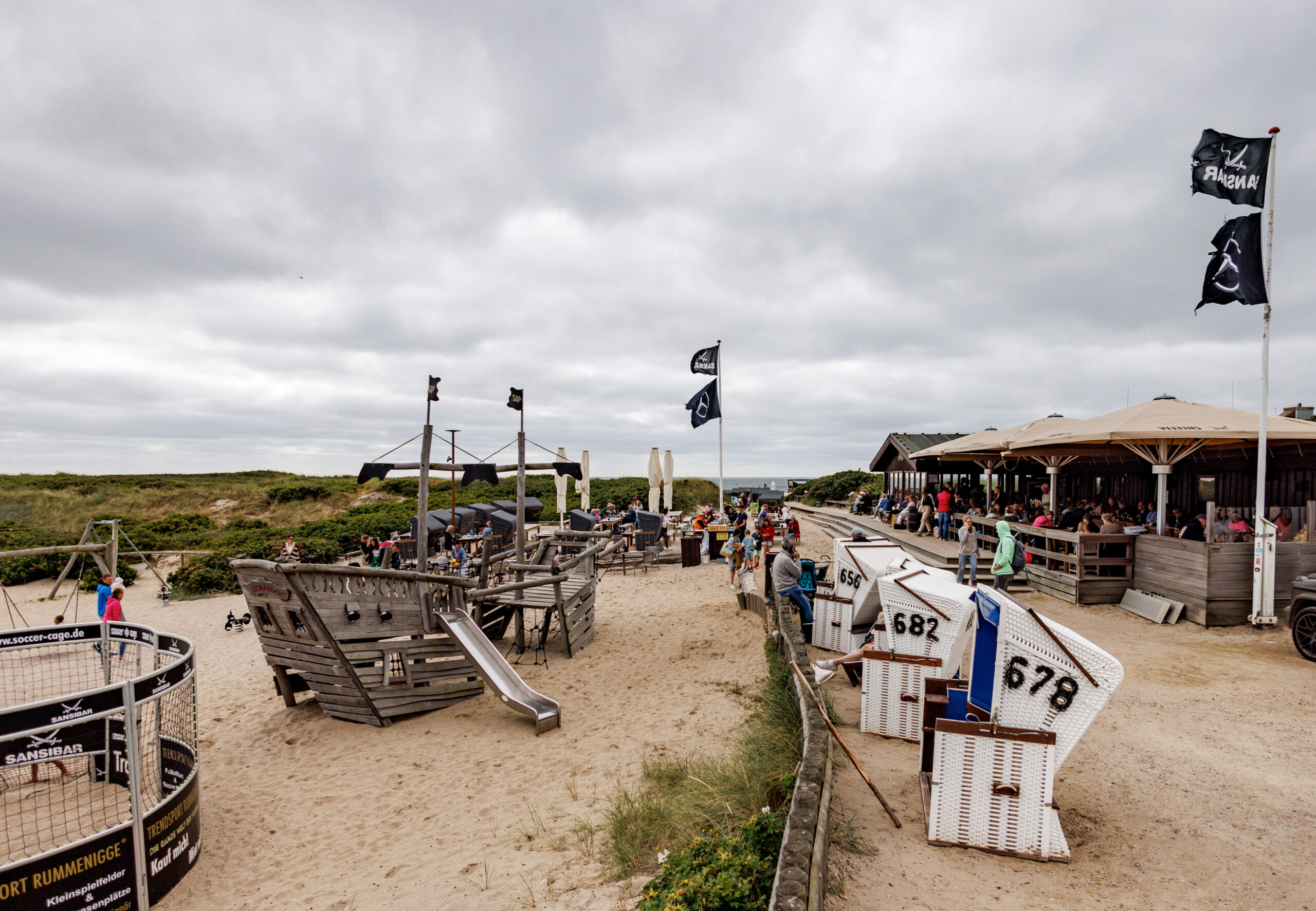 Wolken ziehen über das Restaurant „Sansibar“ auf Sylt. Einige Gäste sitzen draußen. Vorne rechts stehen Strandkörbe, links steht ein Spielplatz. (Archivbild)
