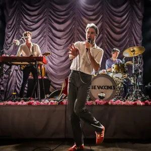 Sänger tänzelnd und singend vorne, dahinter vor dem Vorhang Schlagzeuger und Keyboarder