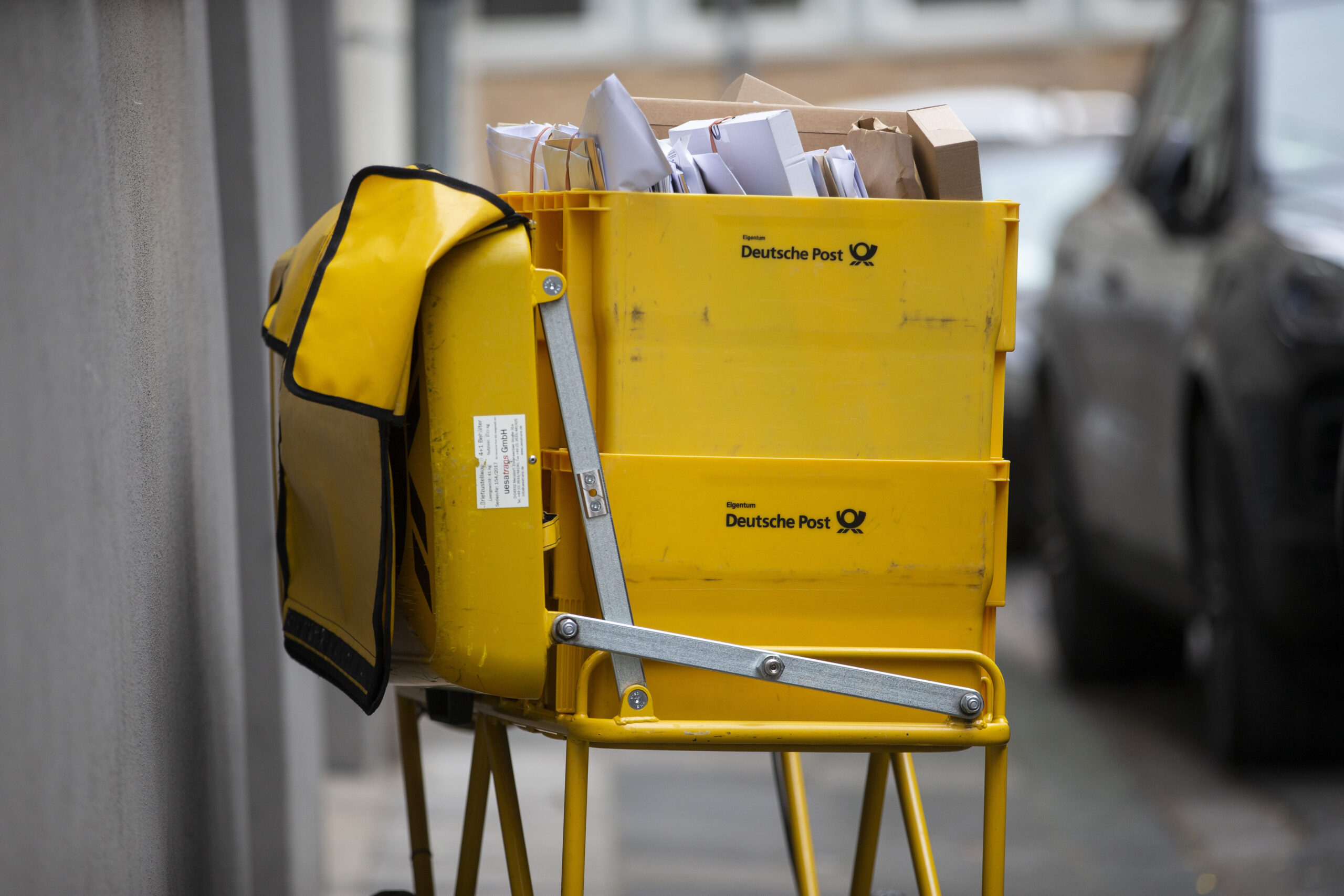 Im Hintergrund eine Straße, im Vordergrund zwei gelbe Post-Kisten voll mit Briefen. Daran hängt links eine gelbe Posttasche