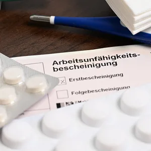 Ein Fieberthermometer, Medikamente und eine ärztliche Arbeitsunfähigkeitsbescheinigung liegen auf einem Tisch. (Symbolbild)