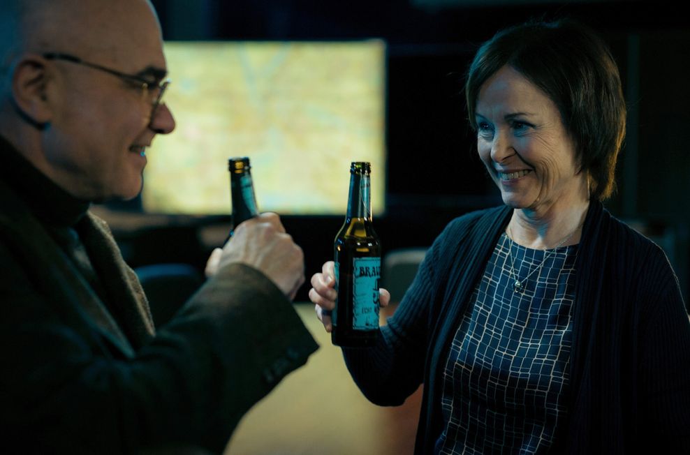 Annalena Schmidt (Edith Keller, r.) und Peter Espeloer (Peter Becker, l.) prosten sich in einer Szene des „Tatort“-Films „Avatar“ mit Bierflaschen in der Hand gegenüberstehend zu.