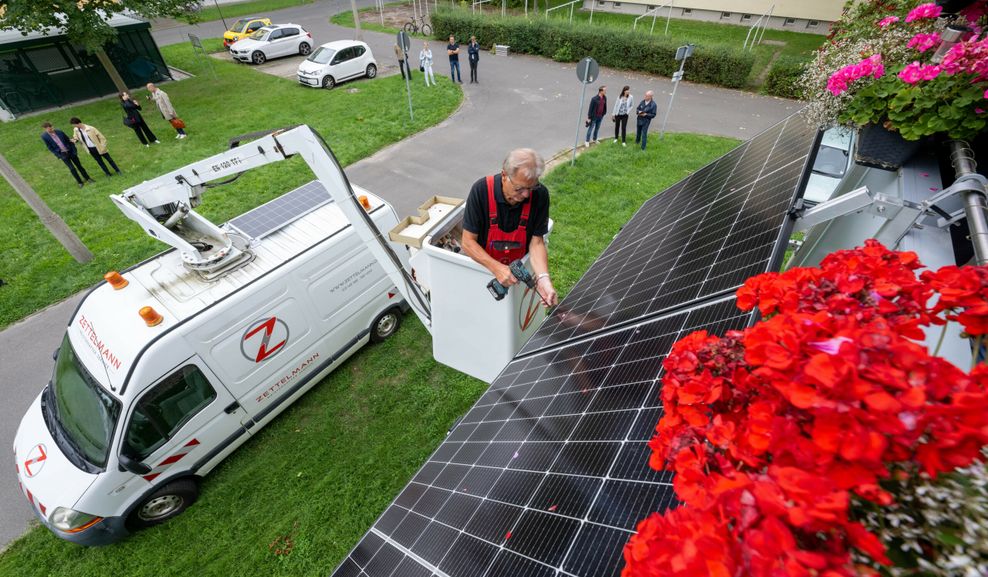 Ein Monteur bringt eine kleine Photovoltaikanlage an einem Balkon an. Im Hintergrund stehen Menschen und schauen zu. (Symbolbild)