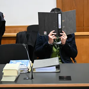 Ein Mann ist in einem Gerichtssaal und verdeckt sich sein Gesicht mit einem Ordner