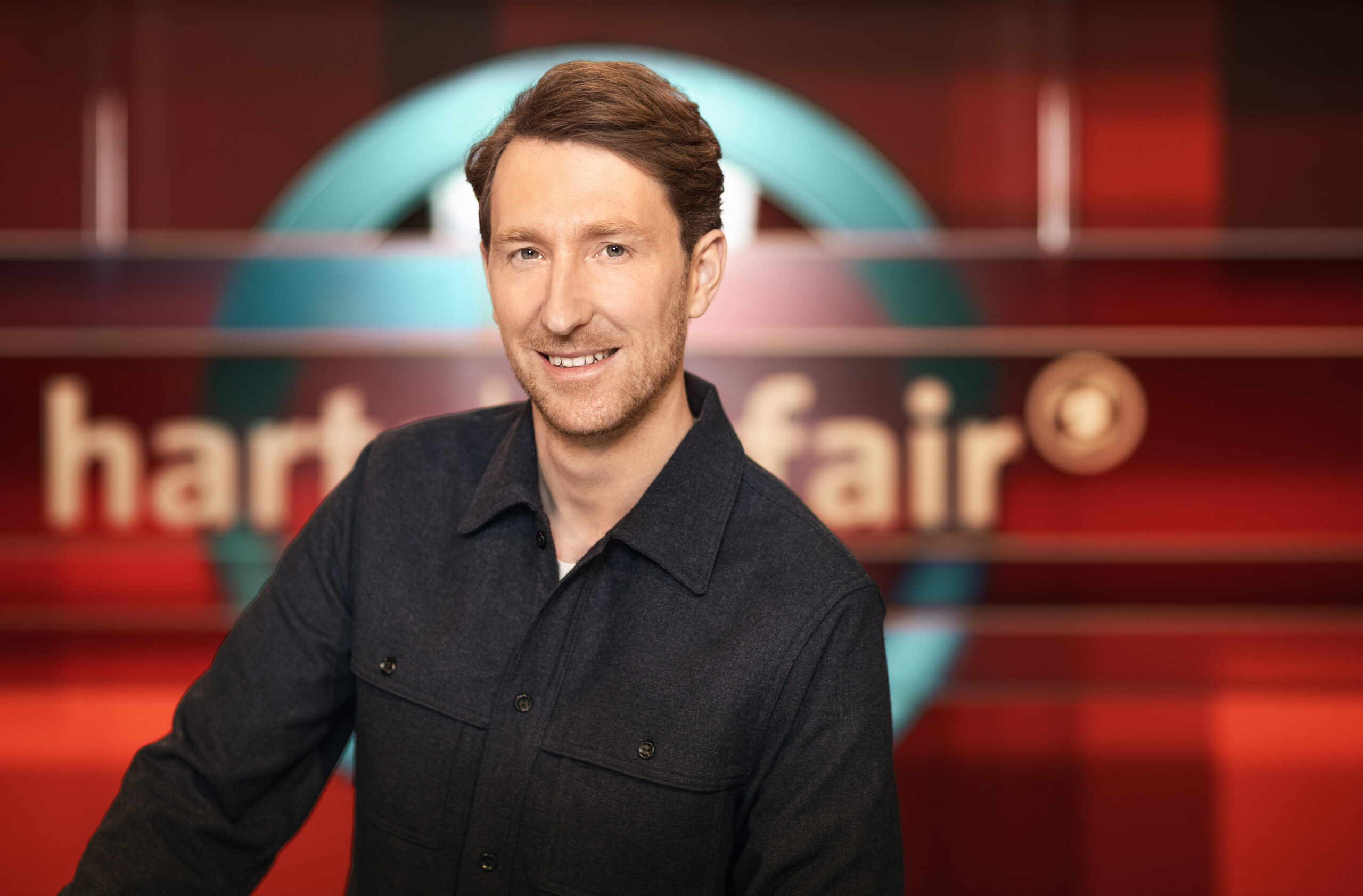 Moderator Louis Klamroth steht vor dem Logo der ARD-Talkshow „Hart aber fair“.