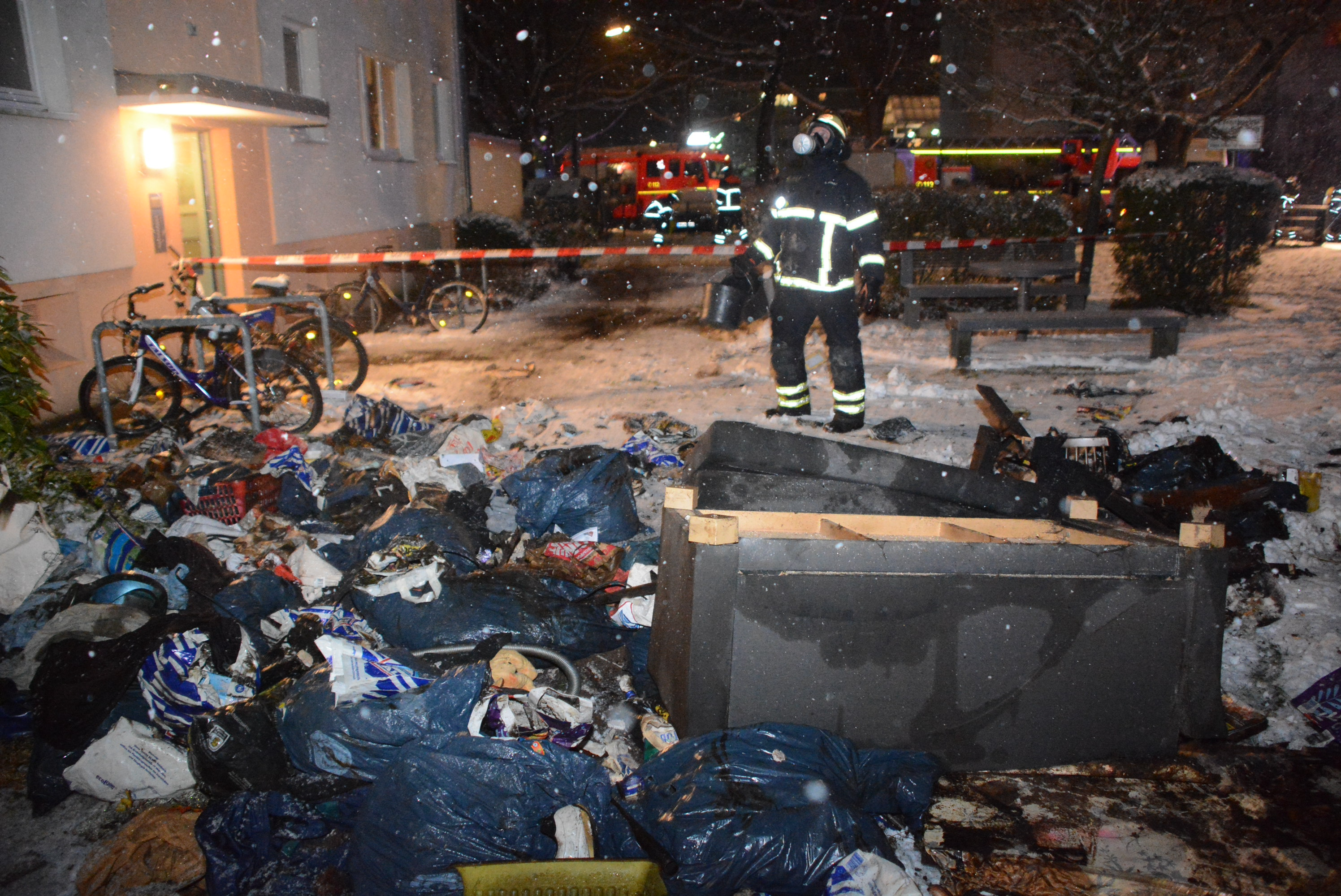 Ein Feuerwehrmann steht vor einem Mehrfamilienhaus und schaut nach oben. Im Vordergrund liegen zahlreiche gefüllte Müllsäcke im Schnee auf dem Boden.