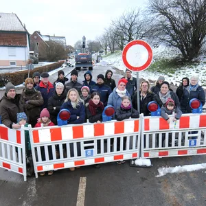 Ca. 25 Anwohner und Anwohnerinnen stehen hinter einer Absperrung, die mit einem Stoppschild die Straße sperrt. Im Hintergrund ein einsamer Bagger