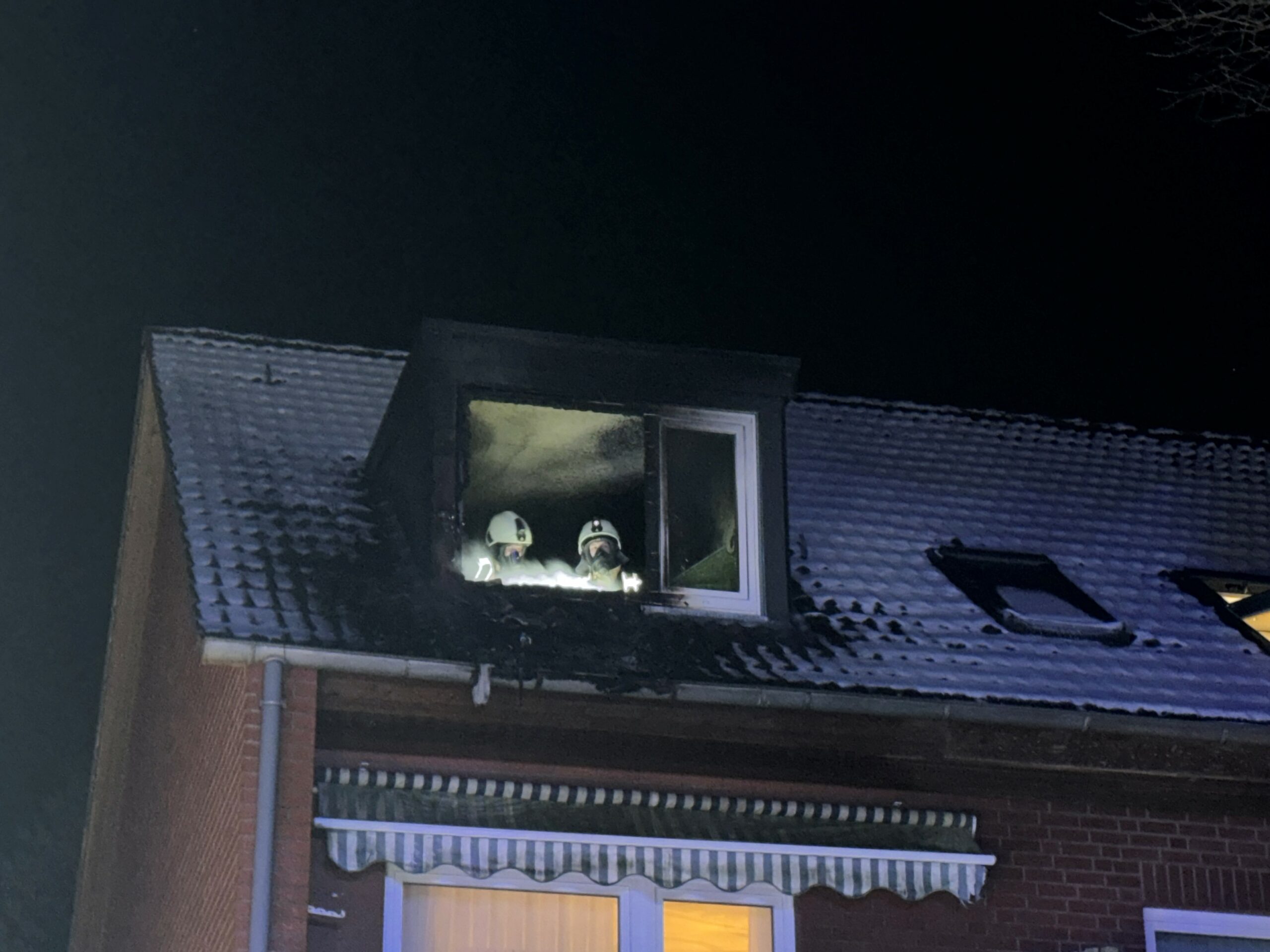 Feuerwehrleute stehen in einer ausgebrannten Wohnung im Dachgeschoss eines Mehrfamilienhauses in Wedel. Sie schauen aus einem Fensterrahmen.