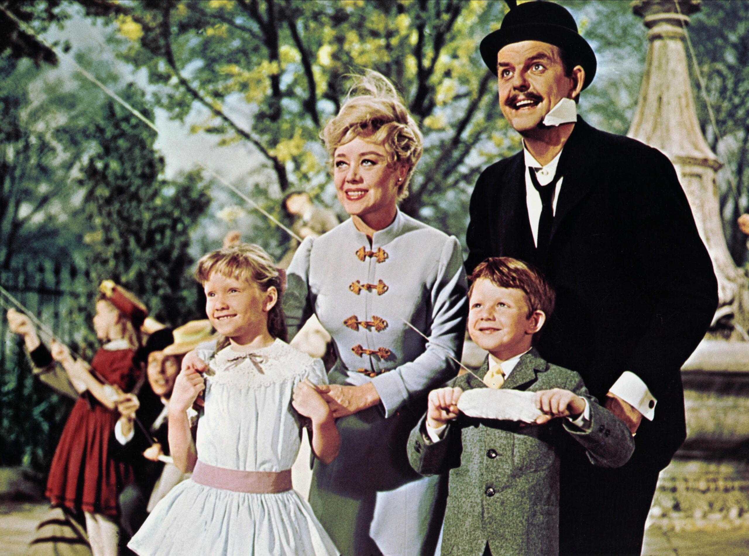 Eine Szene aus dem Disney-FIlm „Mary Poppins“: Karen Dotrice (v.l.n.r.), Glynis Johns, David Tomlinson und Matthew Garber sind in ihren Filmrollen zu sehen. Sie stehen draußen in einem Park.