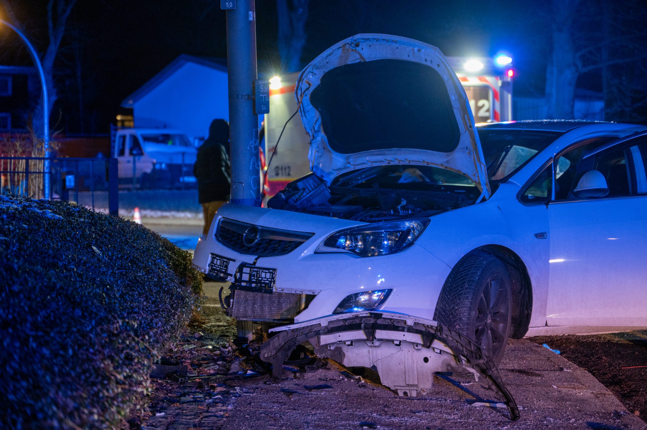 Ein weißes auto steht nach einem Unfall beschädigt an einem Laternenmast. Die Motorhaube steht noch. Im Hintergrund ist ein Rettungswagen zu sehen.