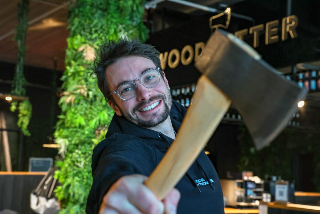 Der Gründer von WoodCutter posiert vor einem WoodCutter Logo mit einer Axt. Er lächelt in die Kamera.