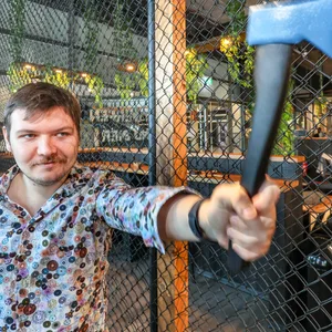Ein junger Mann mit Schnäuzer und Blumen-Hemd wirft eine Axt