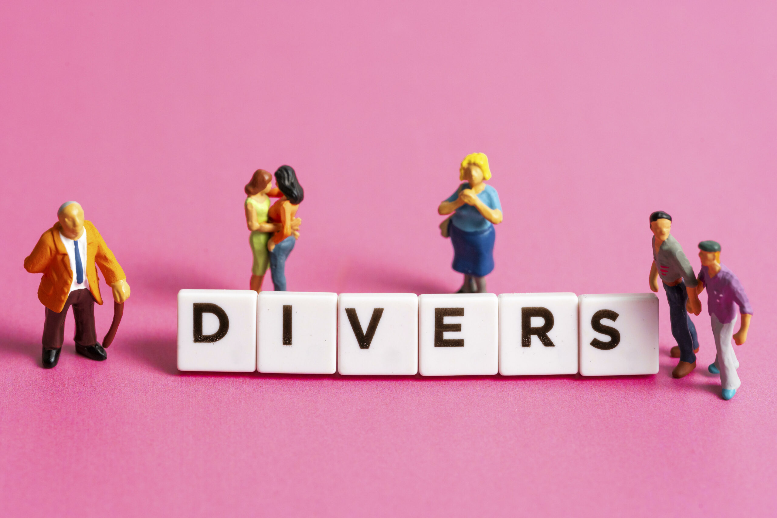 Buchstabenwürfel bilden das Wort Divers neben Miniaturfiguren auf pinkem Hintergrund
