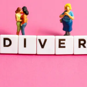 Buchstabenwürfel bilden das Wort Divers neben Miniaturfiguren auf pinkem Hintergrund