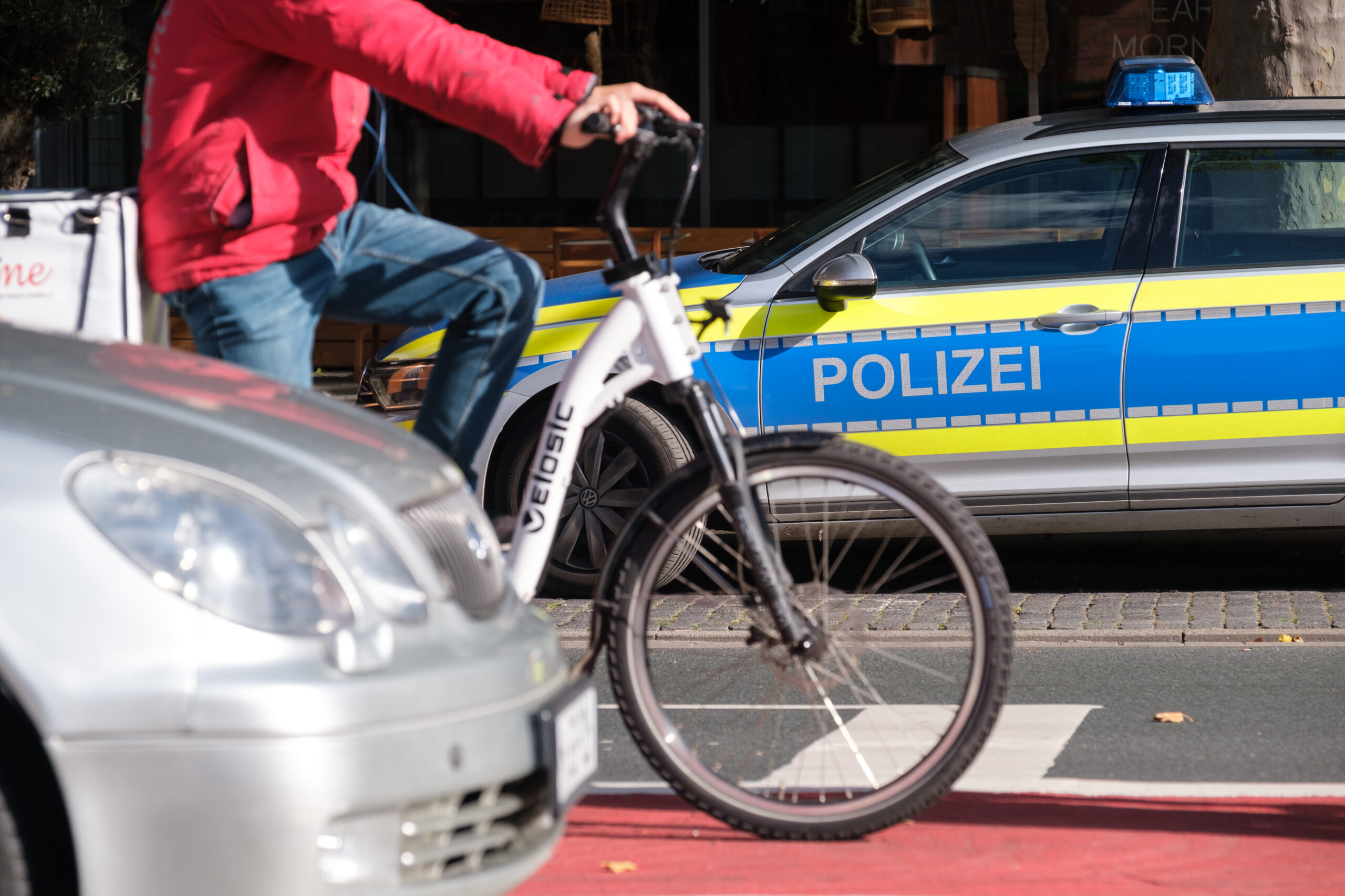 Ein Polizeiauto steht neben einer viel befahrenen Straße, auf der neben einem Auto ein Fahrradfahrer auf einem Radweg hält.