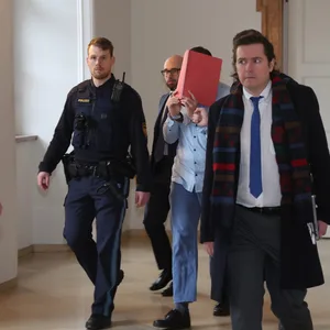Der Angeklagte läuft in den Gerichtssaal. Neben ihm ein Mann im Anzug und ein Justizbeamter. Der geständige Angeklagte hält sich einen roten Ordner vors Gesicht.