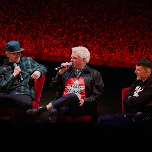 Vier Männer auf der Bühne, sie sitzen auf Sesseln vor einem roten Vorhang, sprechen und lachen