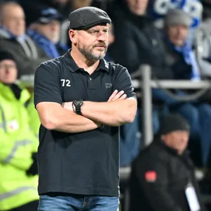 Steffen Baumgart steht mit gekreutzten Armen und skeptischem Blick am Rand des Spielfeldes.