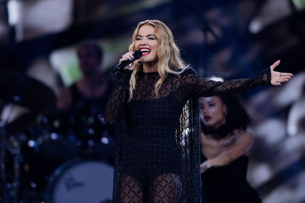 Die Sängerin Rita Ora in einem schwarzen Kleid, ein Mikro in der Hand