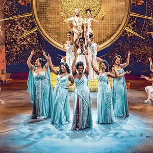 Hercules (Benét Monteiro) im Hintergrund auf der Bühne, davor die Darstellerinnen der Musen in prachtvollen Kleidern